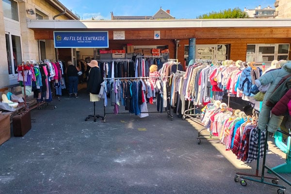Photo de la cour du 23 rue Gouffrand avec les portiques de vêtements et objets divers devant l'entrée de la boutique solidaire au panneau « Au Fil de l'Entraide »